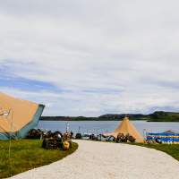 Two tipis sit separately with bunting on shepherds hooks decorating the pathway, the sea and land stand in the background