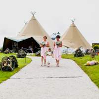 Two bridesmaids and a flower girl walk down a path with three tipis behind them, lobster pots and buoys decorate the path