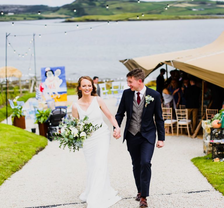 Bride & Groom walk hand in hand, with their wedding Tipi behind them and with an ocean view in background