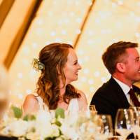 Bride and groom sit and smile, a fairylit panel decorates the inside of the tipi behind them