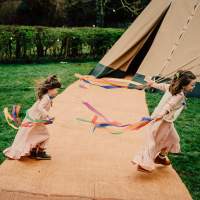 Two girls run with ribbons in their hands in front of a tipi