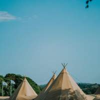 Five tipi tops against a bright blue sky, two blue flags stand in the background against trees