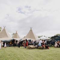Guests gather outdoors and sit on haybales in front of Frankie & Tine's three wedding tipi set up