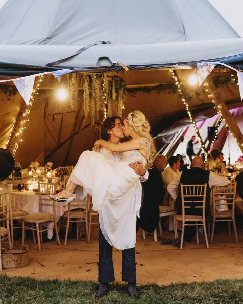 Groom holds the bride as they kiss at the entrance of a tipi decorated by bunting and Fox paper maché of themselves