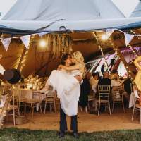 Groom holds the bride as they kiss at the entrance of a tipi decorated by bunting and Fox paper maché of themselves