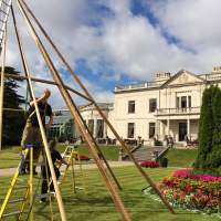Two magnakata crew set up the poles of a tipi, a white mansion with lovely green gardens are in the background