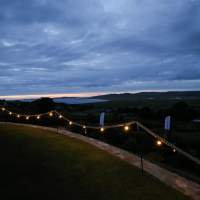 Festoon lighting on ropes are hung on Shepherd's Hooks to decorate the outside of a corporate event
