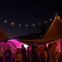 Two Tipis are lit up with festoon lighting and purple lights inside showing corporate guests at night