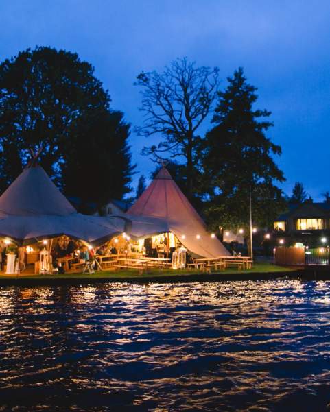 Two tipis are ready for a party, setup on land beside a lake and lit up with fairylights
