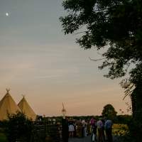 Three tipis stand in the distance with guests and trees in the foreground during sunset