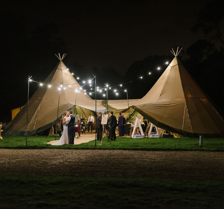 Bride & groom kiss under festoon lighting at night in the entrance of their tipi wedding reception