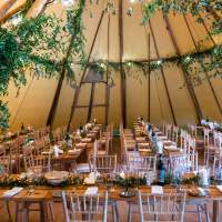 Inside a Tipi is decorated with tables and chairs, lush green foliage on the beam legs framing either side